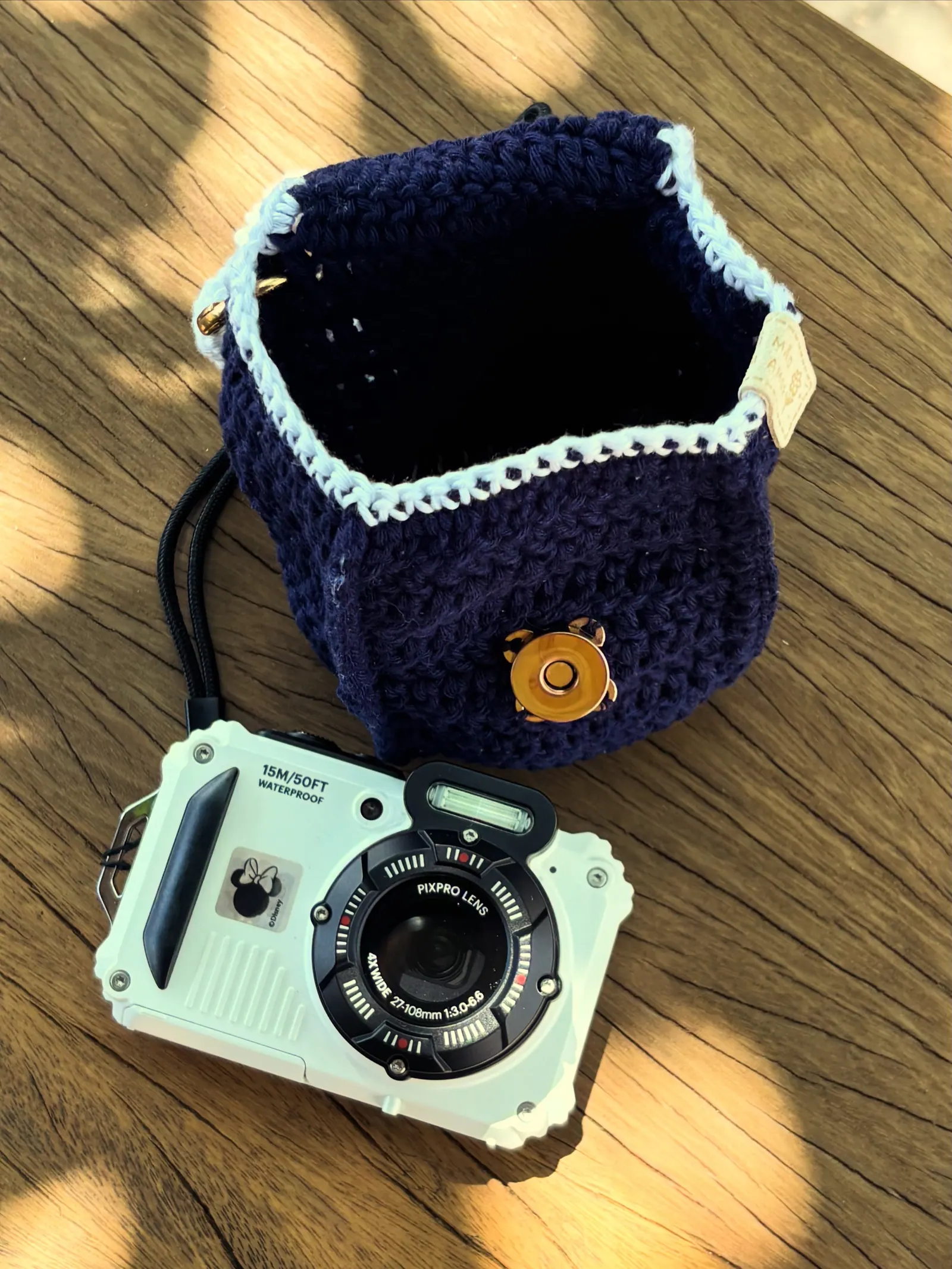Bolsa balde de crochê azul-marinho com borda azul-clara e fecho dourado, ao lado de uma câmera