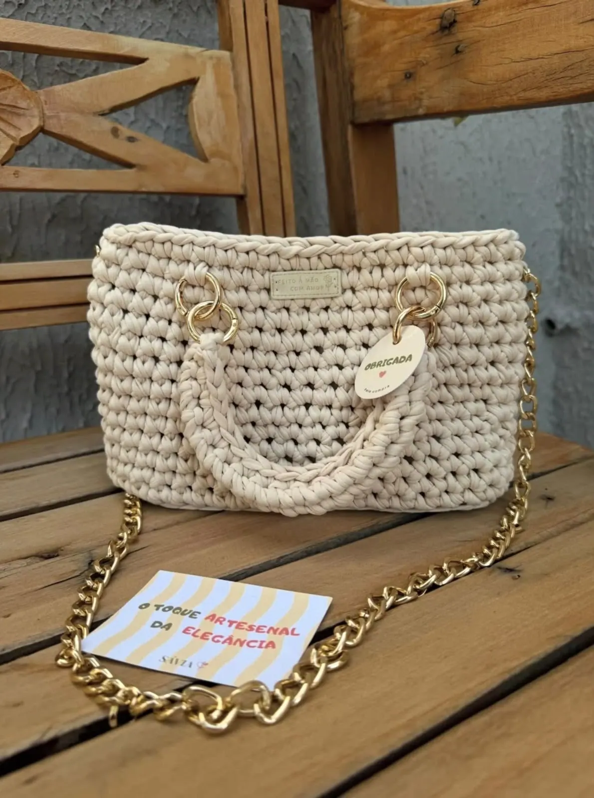 Bolsa de crochê creme com alça de mão e corrente, sobre banco de madeira