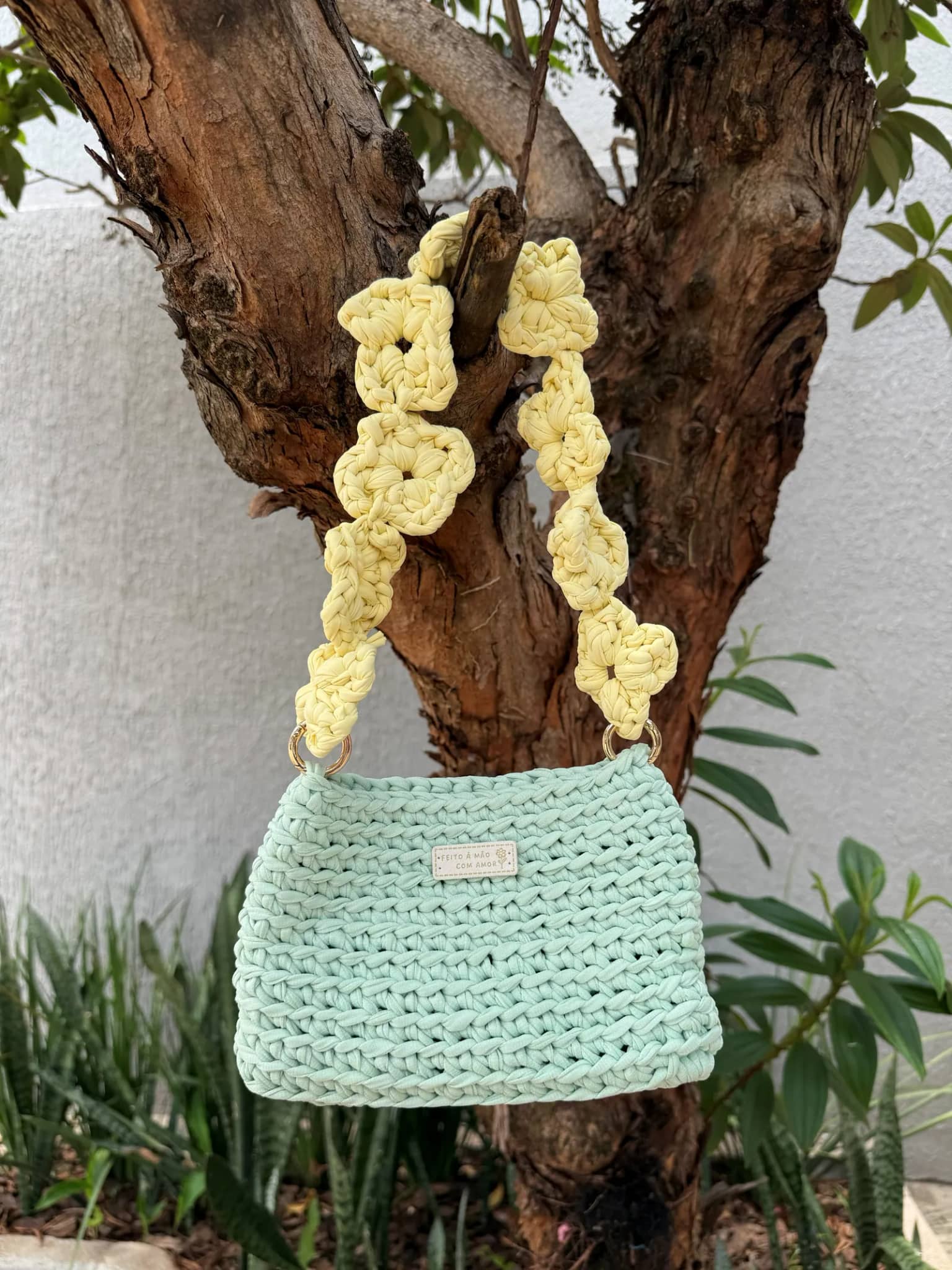 Bolsa de crochê verde-menta com alça de florzinhas amarelas, pendurada em uma árvore