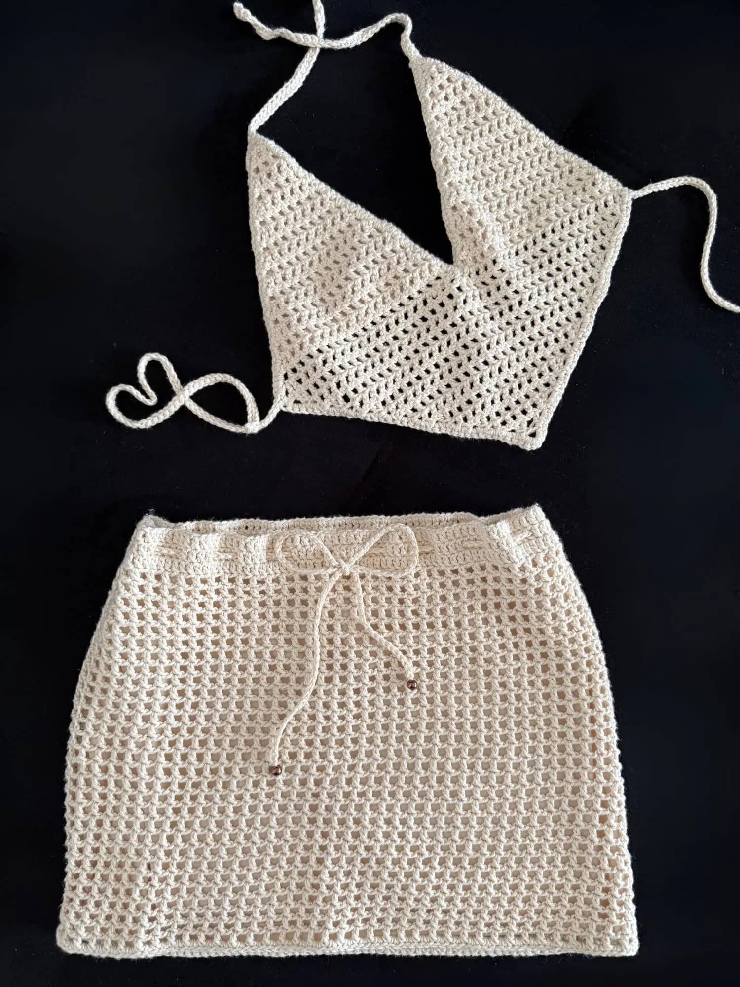 Conjunto de crochê cru com cropped em V e saia curta de trama aberta, sobre fundo preto