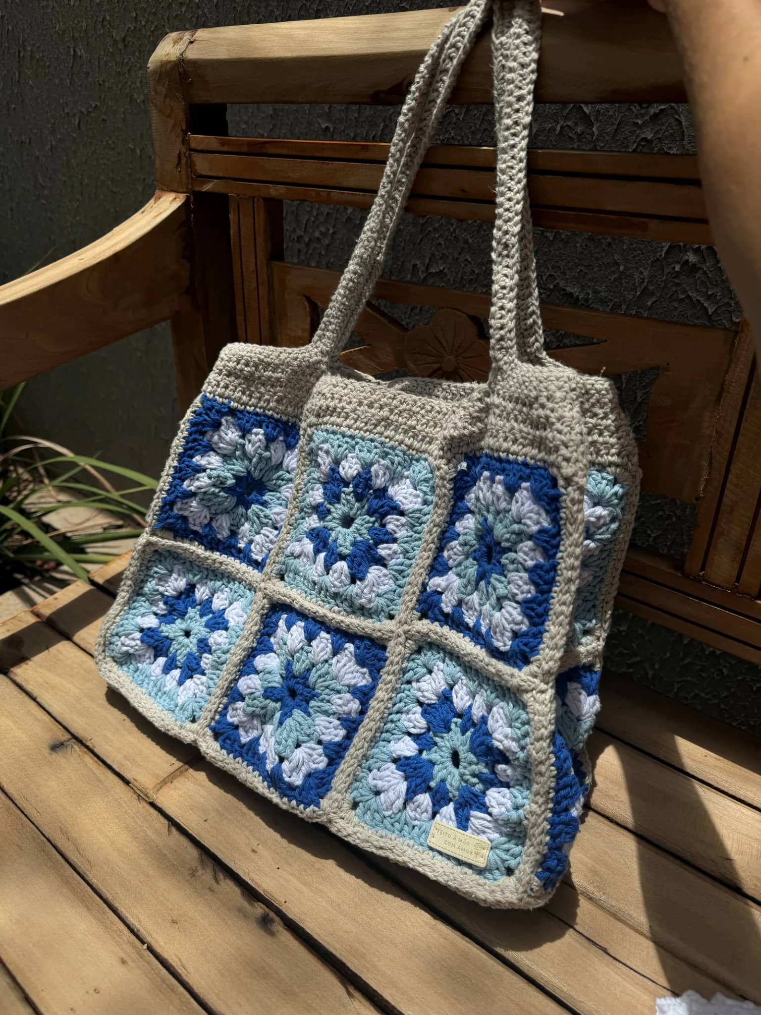 Bolsa tote de crochê com squares azuis e brancos, alças longas bege, sobre um banco de madeira